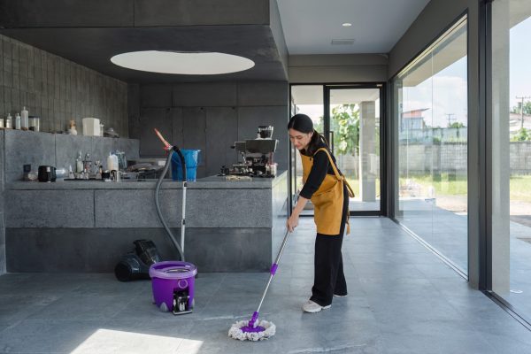 A female employee in a modern coffee shop cleans the floor with a mop and bucket, ensuring a tidy and welcoming environment.