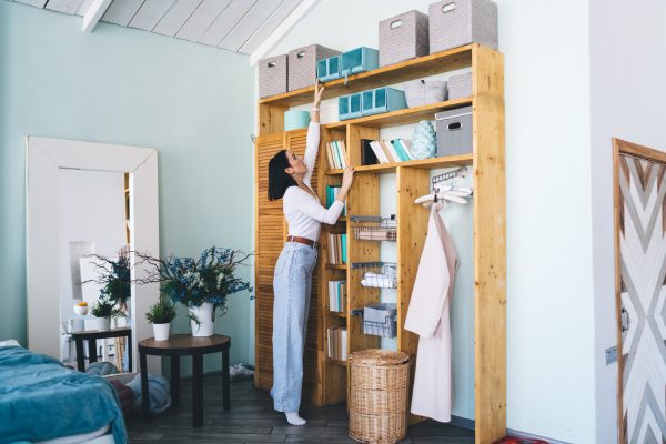 Full body of woman with dark hair in casual wear pulling box from top shelf while doing housework in modern apartment
